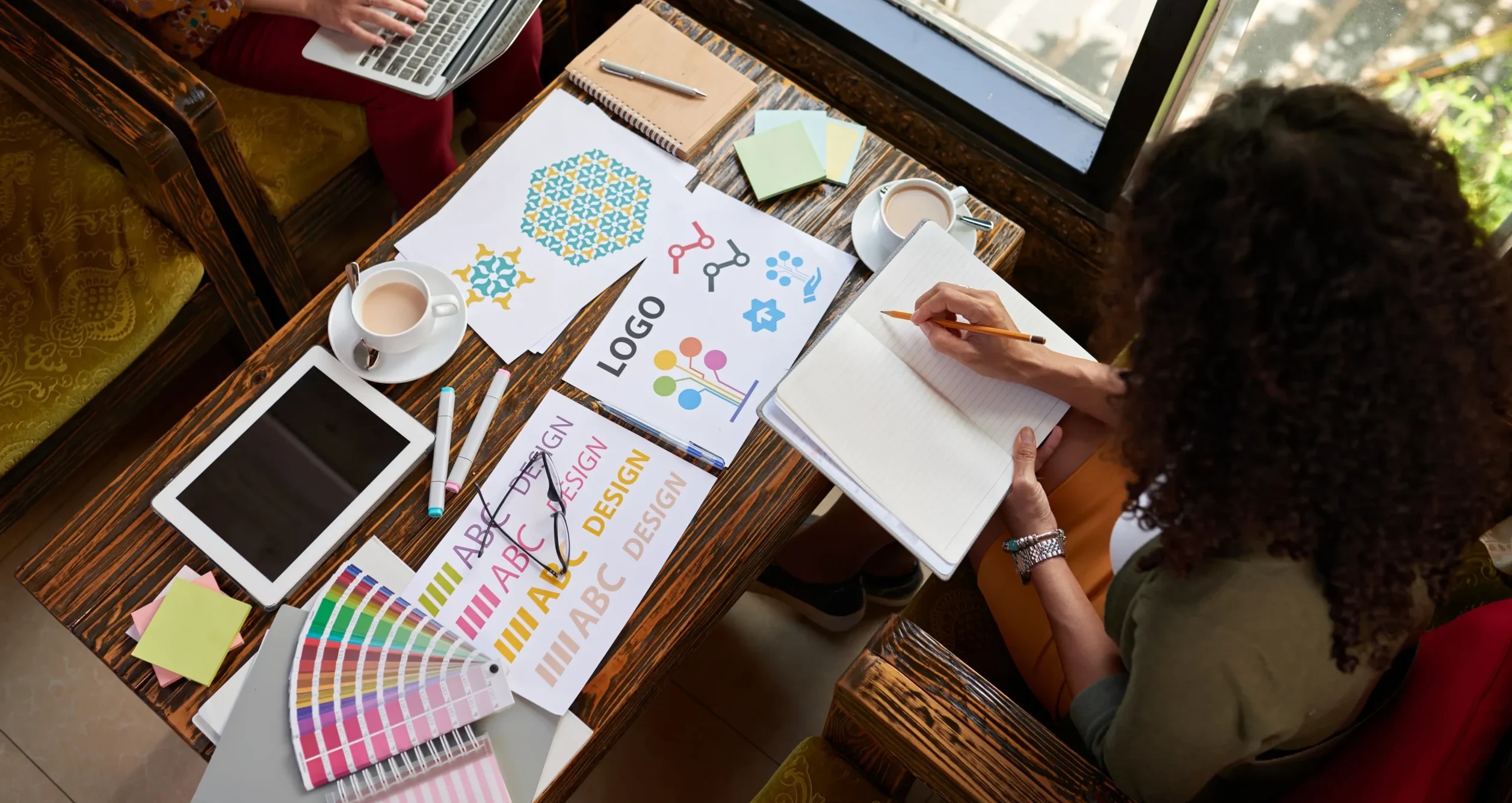 Pessoas trabalhando na criação de uma marca, com esboços de logo, paleta de cores e notebook sobre a mesa.